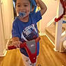 blue_shirt, child, garland, happy, helmet, home, indoor, pacifier, person, playtime, railing, safety_gear, scooter, smiling, stairs, toddler, toy, white_pants, wooden_floor, young_child