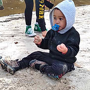 Jayden is registered to the contest to win money with this photo: black_clothing, casual, child, curious, cute, daylight, green_sneakers, hoodie, nature, outdoor, pacifier, person, playing, river, sand, shoes, sitting, toddler, water, young_child