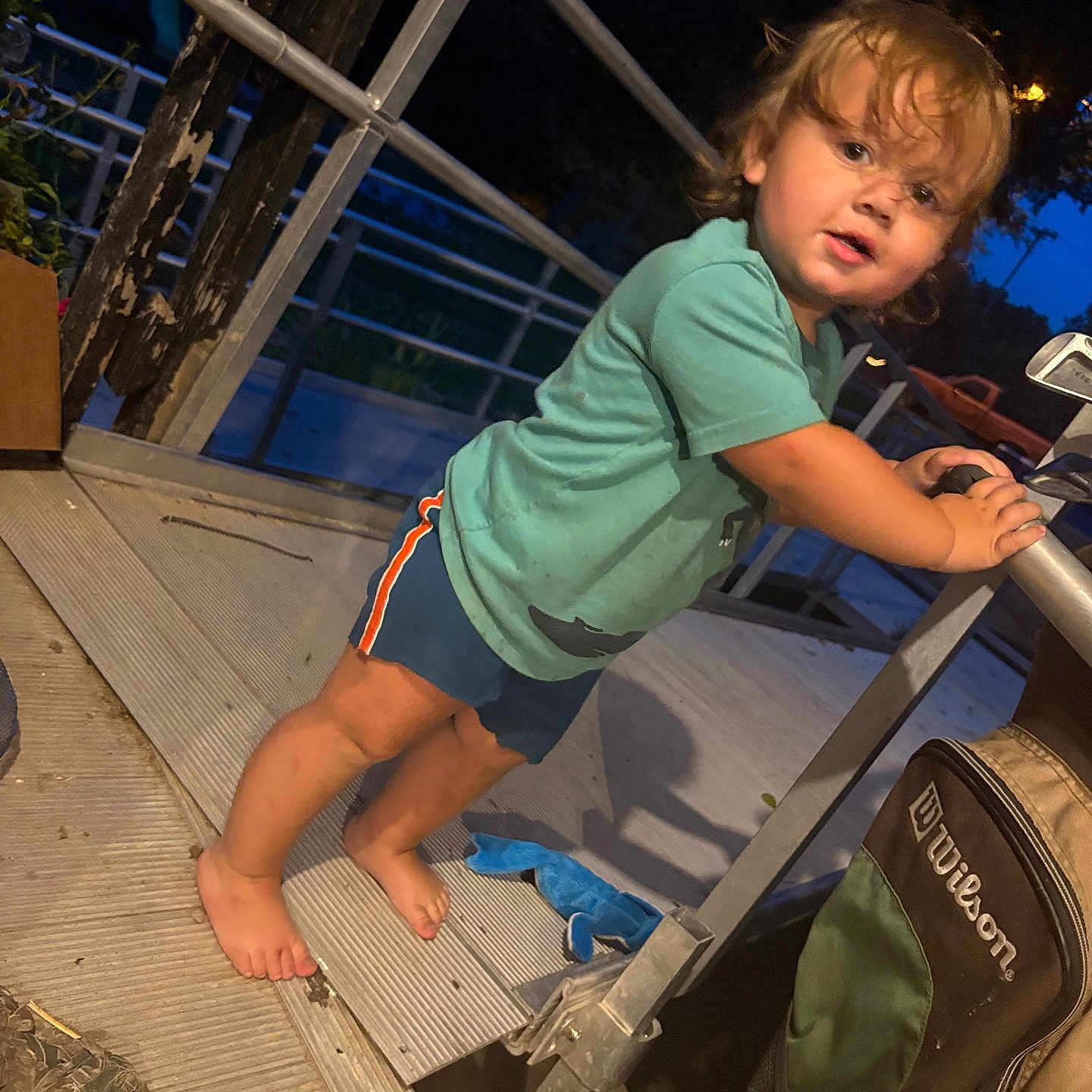 Billy G is registered to the contest to win money with this photo: toddler, child, outdoor, evening, portrait, standing, curious, face, eyes, hair, smiling, green_shirt, shorts, barefoot, stroller, patio, deck, railing, night, candid