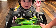 Matthias is registered to the contest to win money with this photo: baby, child, toy_car, camo_hat, indoor, wood_floor, kitchen, cabinet, stove, hand, curious, wide_eyes, green, black, plastic, flooring, playing, person, appliance, expression