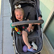 Adalynn is registered to the contest to win money with this photo: baby, stroller, child, smile, toy, foot, bow, happy, outdoor, sidewalk, sunlight, clothing, seatbelt, person, infant, footwear, hand, plastic_toy, fabric, window