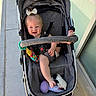 baby, stroller, child, smile, toy, foot, bow, happy, outdoor, sidewalk, sunlight, clothing, seatbelt, person, infant, footwear, hand, plastic_toy, fabric, window