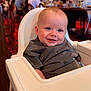 baby, high_chair, child, smiling, drool, grey_shirt, indoor, restaurant, people, blurred_background, wooden_chair, lighting, portrait, happy, infant, face, person, seated, casual_clothing, table