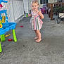 barefoot, casual, child, concrete_floor, curious_expression, daylight, door, home, outdoor, person, plant, plastic_toy, playful, porch, railing, small_hair_ties, striped_dress, toddler, toys, wooden_chair