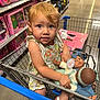 toddler, child, shopping_cart, doll, toy_store, aisle, toys, floral_dress, blue_eyes, shopping, plastic, retail, store_shelves, floor, person, holding, cute, indoor, playful, curious