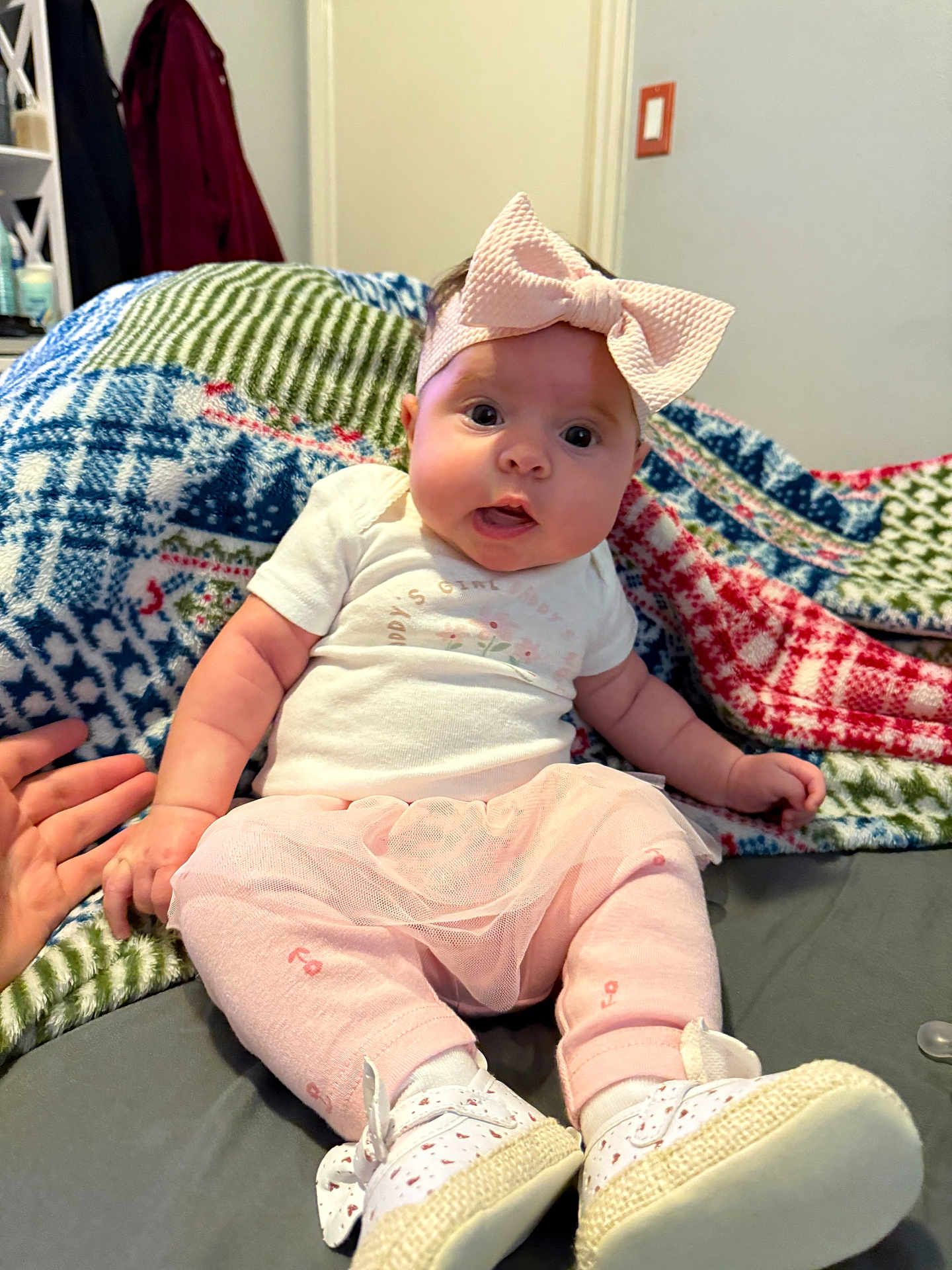 Karlina M Yokley is registered to the contest to win money with this photo: baby, child, infant, tutu, headband, pink_clothing, hand_holding, blanket, patterned_blanket, sitting, footwear, white_shoes, indoor, person, cute, expression, face, wide_eyes, soft_lighting, home