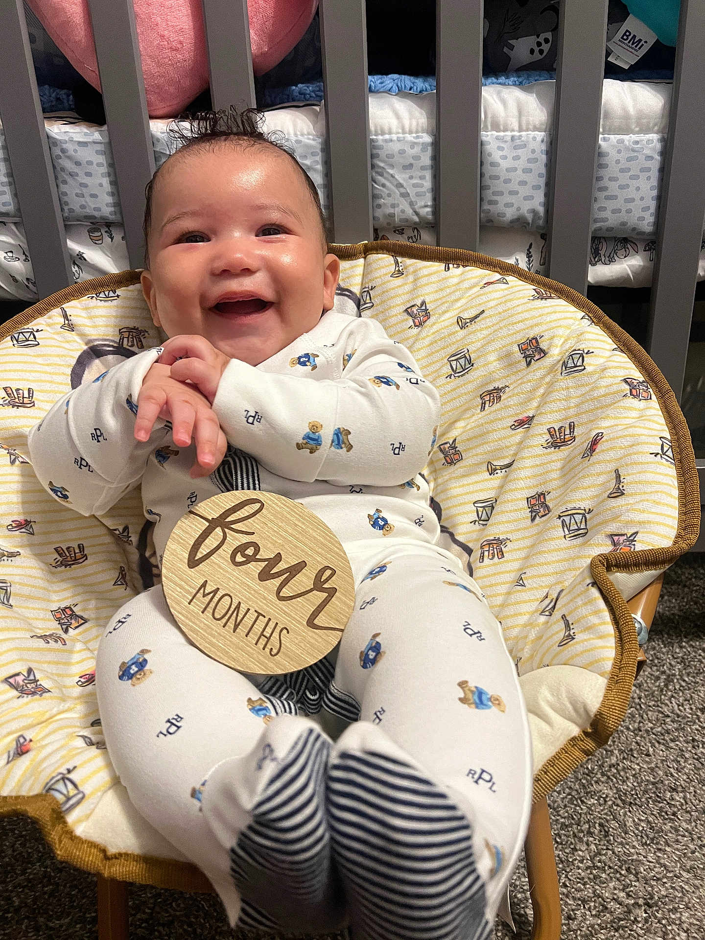 Jrue is registered to the contest to win money with this photo: baby, infant, smile, happy, onesie, chair, patterned, clothing, hands, feet, indoor, carpet, crib, wood, fabric, cute, child, person, young, four_months