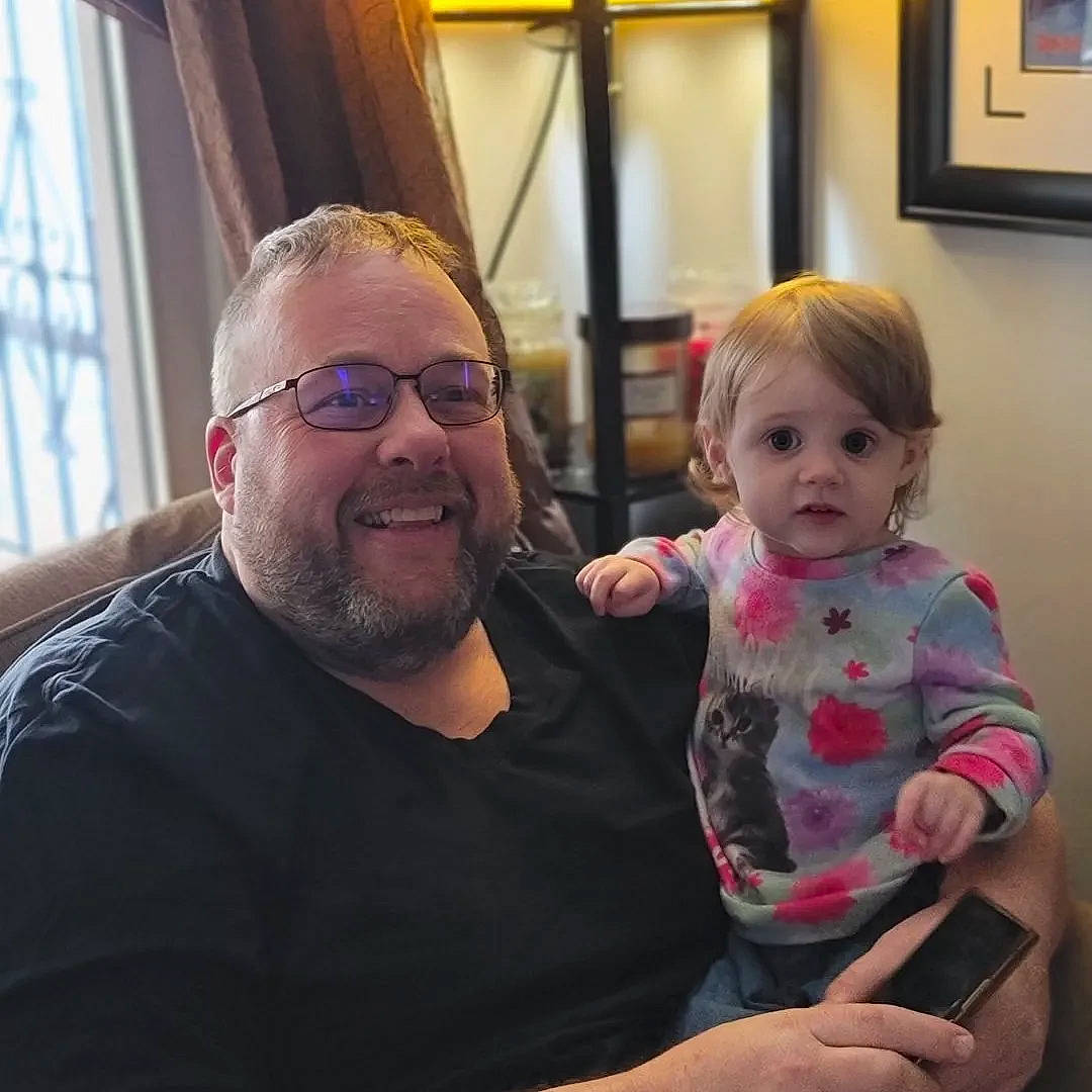 Juno is registered to the contest to win money with this photo: baby, beard, chin, comfort, curtain, event, eyewear, fun, glasses, happy, human_body, iris, joy, lap, person, picture_frame, shoulder, smile, t_shirt, toddler