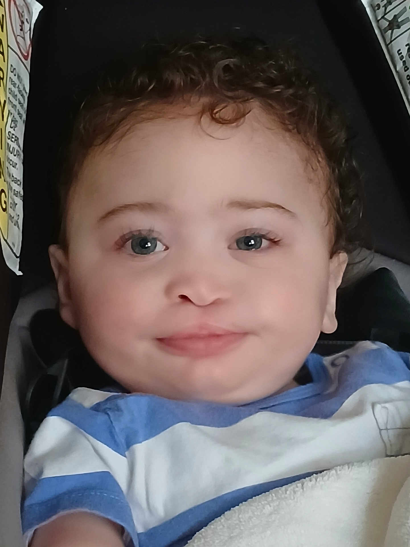 Elijah is registered to the contest to win money with this photo: baby, child, curly_hair, blue_eyes, striped_shirt, infant, portrait, close_up, car_seat, blanket, smile, face, young_child, cute, person, indoors, comfort, young_baby, seated, casual_clothing