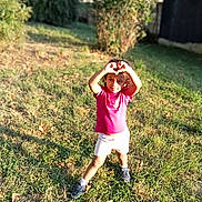 Natalia participe au concours pour gagner de l'argent avec cette photo : child, grass, outdoor, sunlight, greenery, shirt, shorts, sneakers, hands, heart_shape, standing, nature, daylight, happy, person, curly_hair, playing, summer, casual_clothing, park