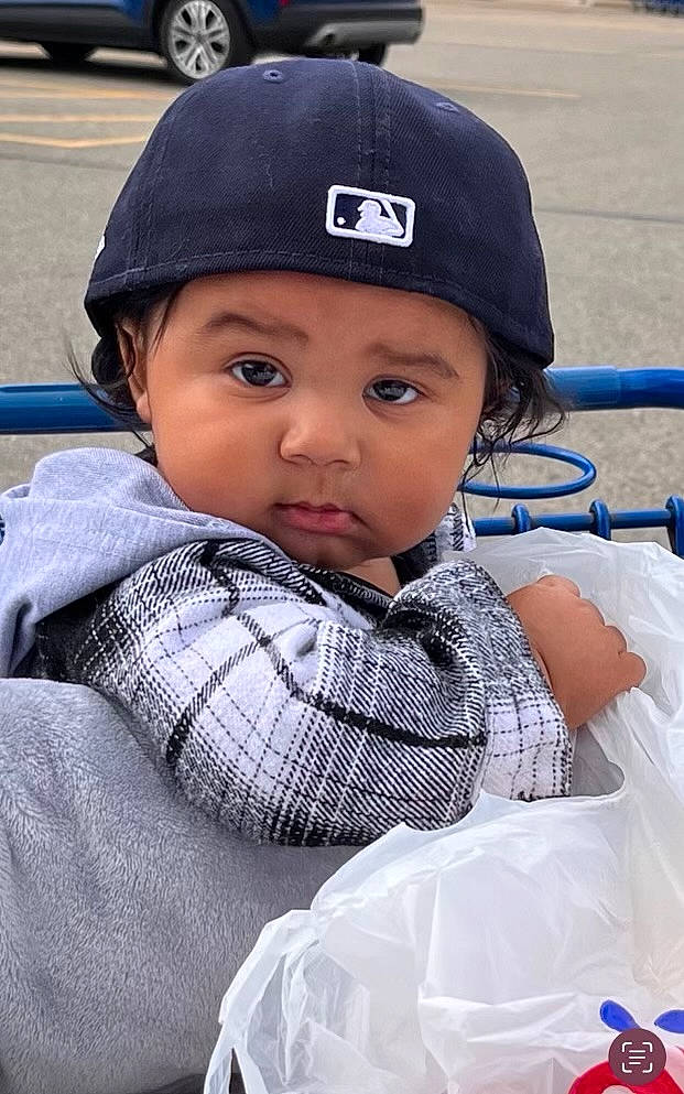 Tre is registered to the contest to win money with this photo: baseball_cap, blue, cap, child, electric_blue, event, fashion_accessory, fun, headwear, pattern, people, person, personal_protective_equipment, plaid, plastic_bag, recreation, sitting, toddler, travel, uniform