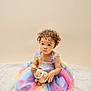 toddler, child, curly_hair, rainbow_dress, plush_toy, bunny, sitting, blanket, soft_texture, indoor, portrait, cute, colorful_clothing, baby, innocent, expression, studio_lighting, pastel_colors, young_child, background