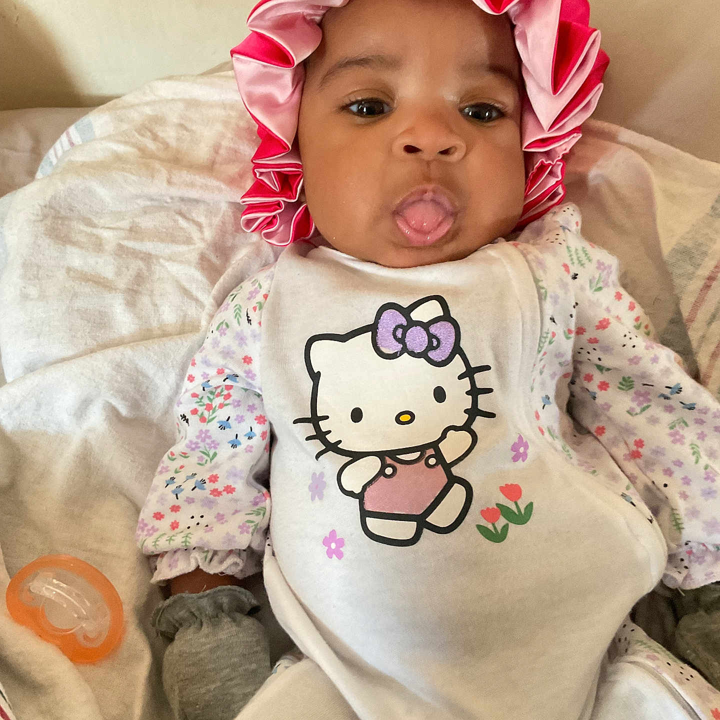 Suraaya joined the competition — help win amazing prizes! baby, blanket, child, clothing, cozy, cute, floral_pattern, hello_kitty, indoors, infant, laying_down, mittens, pacifier, person, pink_bonnet, playful, portrait, smiling, soft_lighting, tongue_out