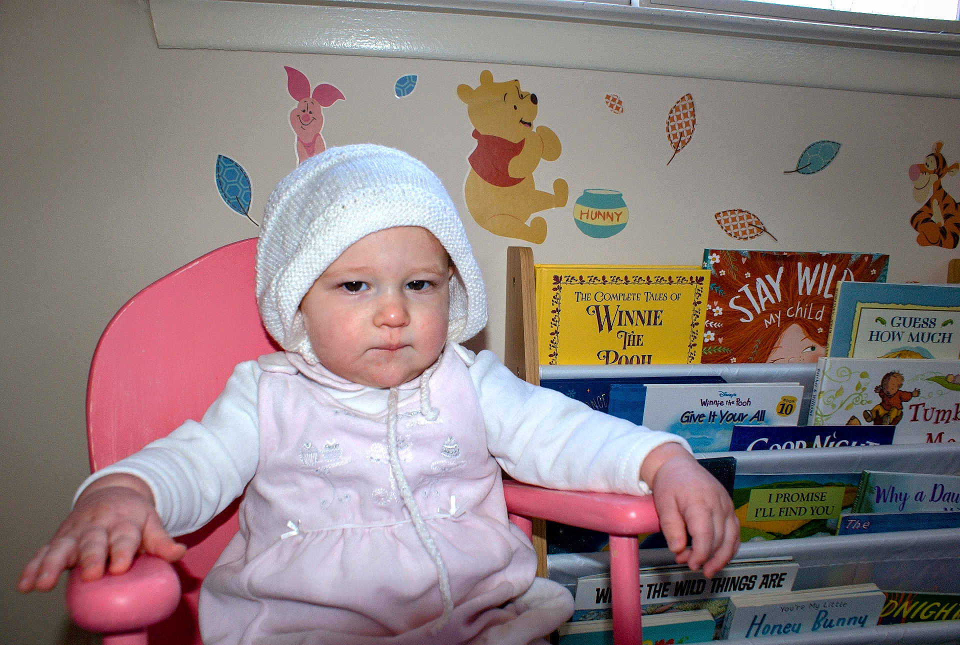 Indigo joined the competition — help win amazing prizes! child, baby, toddler, pink_chair, knitted_hat, book_shelf, books, winnie_the_pooh, piglet, tigger, wall_decals, nursery, grumpy_expression, portrait, hand, arm, white_clothing, indoor, seat, picture_books