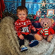 Luka is registered to the contest to win money with this photo: toddler, child, smiling, sweater, mickey_mouse, teddy_bear, christmas, holiday, decorations, chair, fluffy, shoes, indoors, portrait, festive, winter, pine_trees, scarf, red, cozy