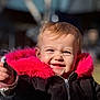 child, toddler, baby, smiling, toothy_grin, pink_fur, hooded_jacket, black_coat, close_up, portrait, outdoor, winter_clothing, hand, zipper, sunlight, rosy_cheeks, shallow_depth_of_field, happy, playground, blonde_hair