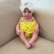 Blake is registered to the contest to win money with this photo: baby, child, infant, bow, headband, yellow_clothing, striped, couch, beige, sitting, cute, portrait, indoors, small_feet, smiling, happy, person, furniture, skin, blue_eyes