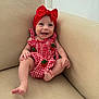 baby, smiling, red_bow, red_dress, strawberry_pattern, couch, beige, happy, child, infant, sitting, indoors, cute, portrait, headwear, feet, hands, skin, person, young_child