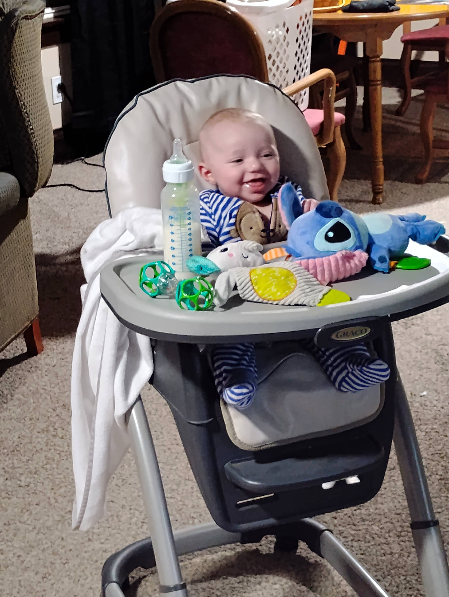 Cyrus is registered to the contest to win money with this photo: baby, blanket, bottle, carpet, chair, child, cute, furniture, happy, high_chair, home, indoor, infant, living_room, person, plush_toy, smile, striped_pajamas, table, toy