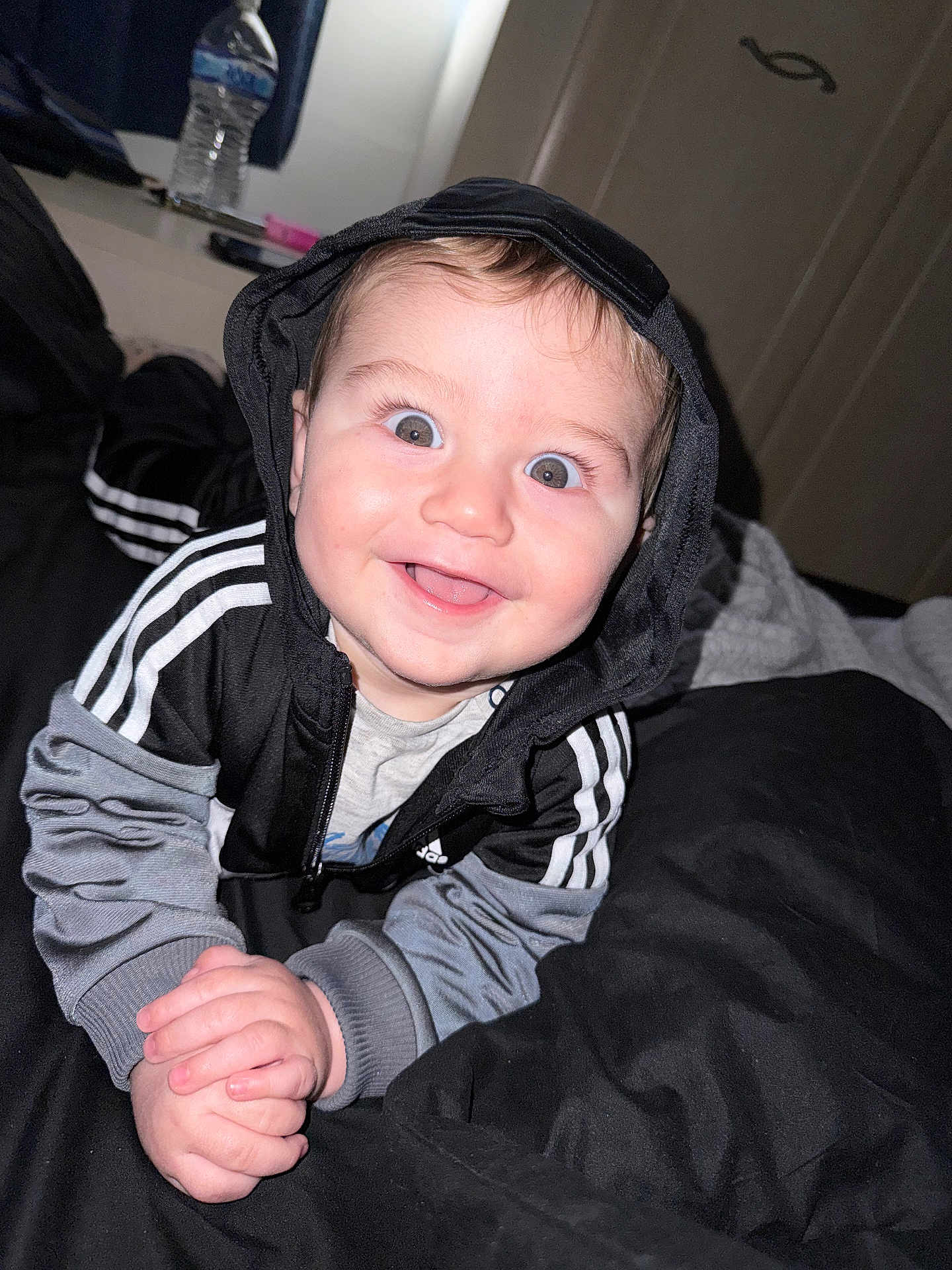 Marsh is registered to the contest to win money with this photo: baby, child, hoodie, black_clothing, gray_clothing, smiling, happy, face, hands, bed, blanket, indoor, person, cute, eyes, mouth, infant, clothing, portrait, baby_face