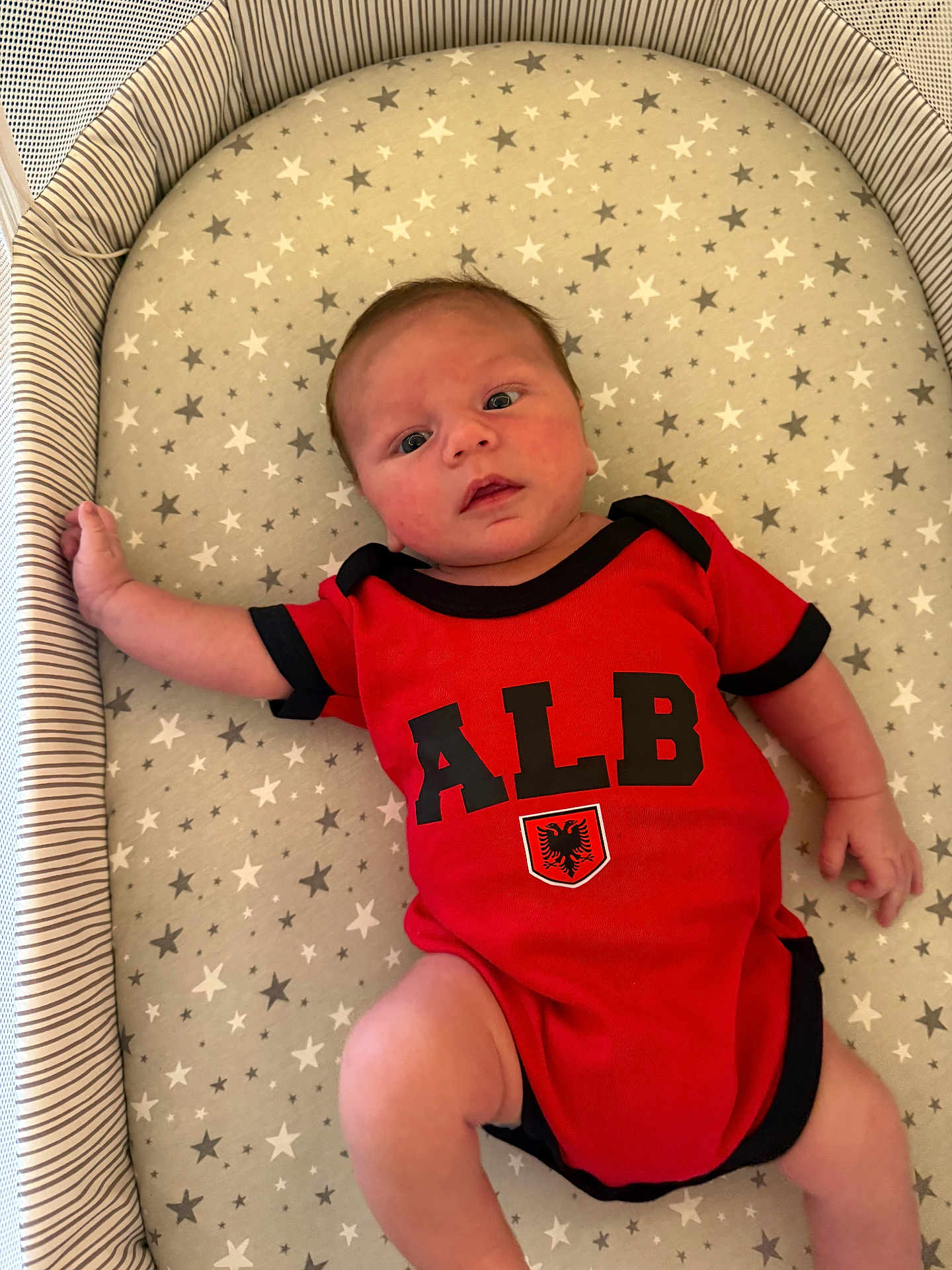 Marsh is registered to the contest to win money with this photo: baby, infant, onesie, red_clothing, face, laying_down, star_pattern, mattress, striped_bed, hand, foot, person, newborn, cute, indoor, soft_lighting, skin, expression, young_child, comfortable