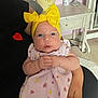 adult_arm, baby, bottle, child, curious_expression, eyes, face, floor, fruit_pattern, furniture, hands, headband, holding, indoor, infant, person, pink_dress, seated, side_table, yellow_bow