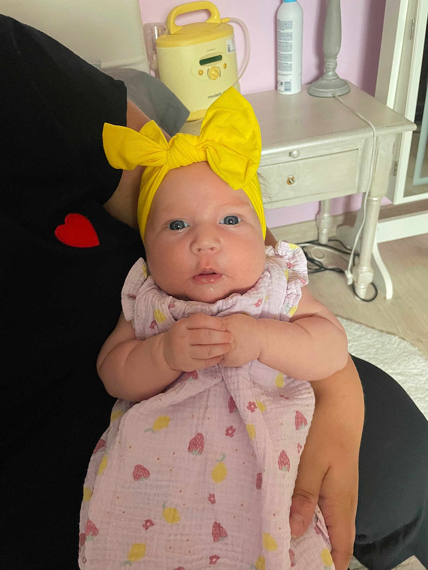 Esmée participe au concours pour gagner de l'argent avec cette photo : adult_arm, baby, bottle, child, curious_expression, eyes, face, floor, fruit_pattern, furniture, hands, headband, holding, indoor, infant, person, pink_dress, seated, side_table, yellow_bow