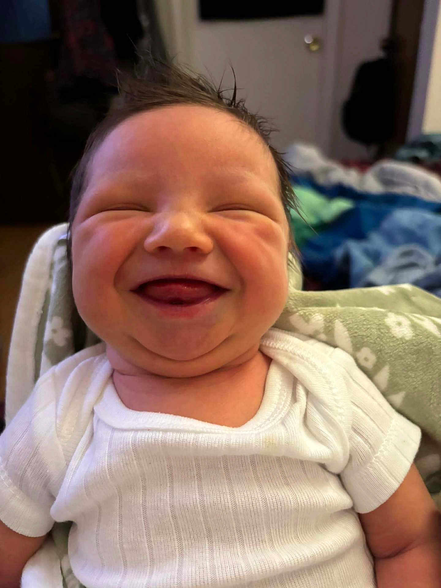 Tatum is registered to the contest to win money with this photo: baby, smiling, happy, infant, face, hair, white_shirt, blanket, cute, expression, indoors, person, child, newborn, portrait, skin, chubby_cheeks, tongue_out, cozy, casual