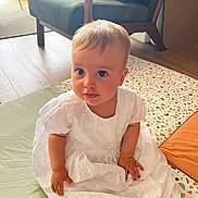 Anaé a rejoint le concours — aidez-le/la à gagner de superbes lots ! baby, child, white_dress, sitting, floor, mat, patterned_mat, indoor, furniture, armchair, wooden_floor, curious, cute, person, infant, natural_light, home, soft_light, looking_up, portrait
