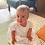baby, child, white_dress, sitting, floor, mat, patterned_mat, indoor, furniture, armchair, wooden_floor, curious, cute, person, infant, natural_light, home, soft_light, looking_up, portrait
