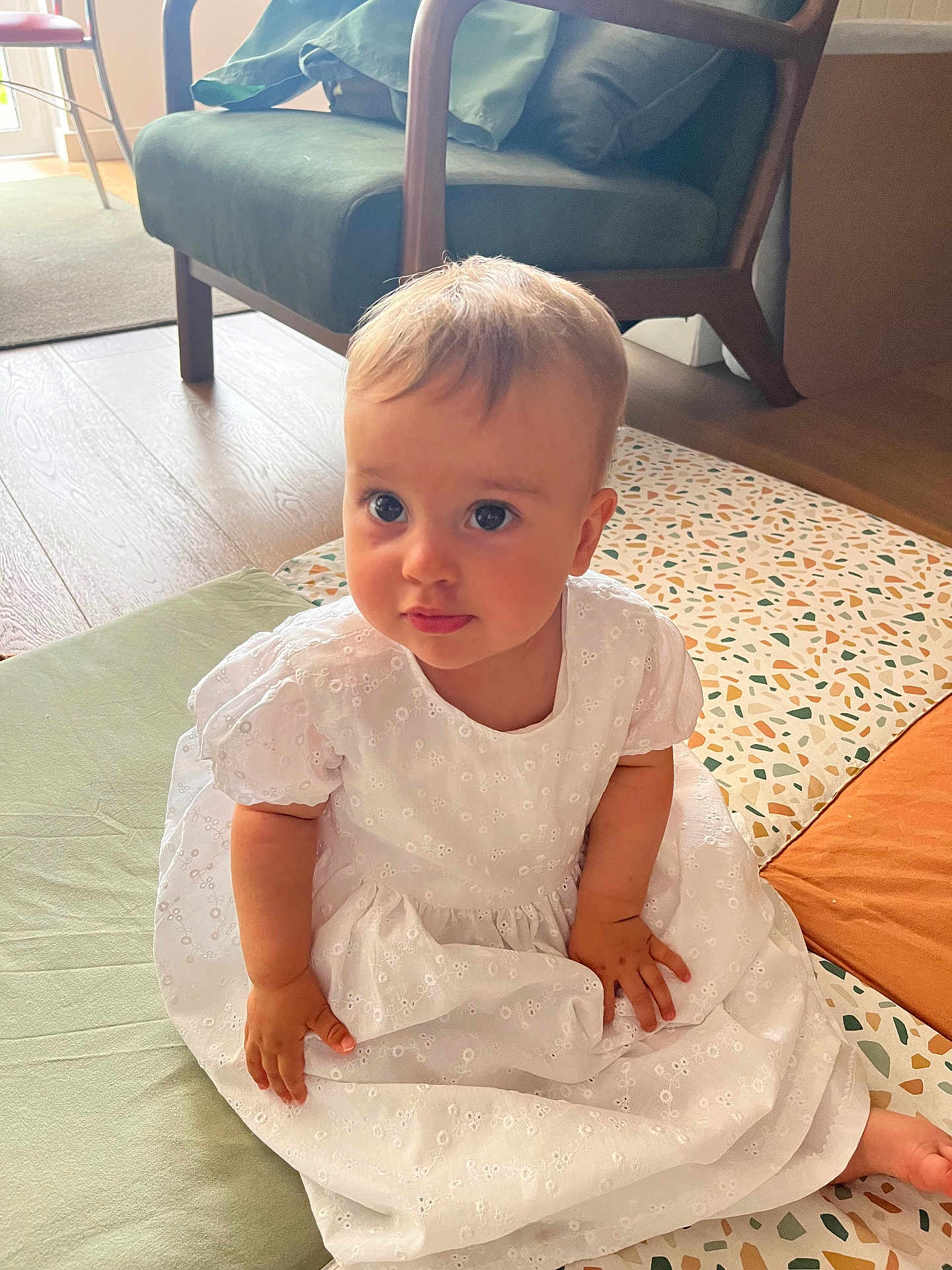 Anaé a rejoint le concours — aidez-le/la à gagner de superbes lots ! baby, child, white_dress, sitting, floor, mat, patterned_mat, indoor, furniture, armchair, wooden_floor, curious, cute, person, infant, natural_light, home, soft_light, looking_up, portrait
