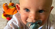 Tommy is registered to the contest to win money with this photo: baby, blue_eyes, carpet, child, close_up, curious, cute, happy, indoor, infant, light, pacifier, person, plush_toy, smiling, soft_texture, toddler, toy, white_clothing, young_child