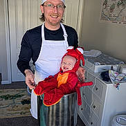Owen is registered to the contest to win money with this photo: man, baby, costume, lobster_costume, chef_hat, apron, pot, smile, indoor, carpet, cabinet, glasses, door, person, child, happy, playful, funny, home, kitchen