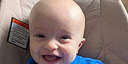 Owen is registered to the contest to win money with this photo: baby, smiling, blue_sweater, bald_head, happy, infant, baby_seat, cushion, indoor, child, face, clothing, portrait, person, seat, warning_label, closeup, cute, young, comfortable