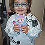 child, glasses, toy, plush_toy, indoor, vacuum_cleaner, pajamas, smiling, hair_ties, floor_tiles, person, cute, young, holding, colorful, standing, face, eyes, hands, sweatshirt
