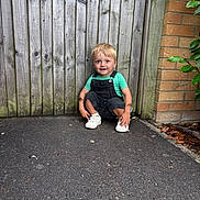 Jackson joined the competition — help win amazing prizes! child, blonde_hair, green_shirt, overalls, white_shoes, squatting, wooden_fence, brick_wall, outdoor, pavement, green_leaves, happy, smiling, tattoos, casual_clothing, daylight, curious, young_child, nature, playful