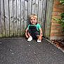 child, blonde_hair, green_shirt, overalls, white_shoes, squatting, wooden_fence, brick_wall, outdoor, pavement, green_leaves, happy, smiling, tattoos, casual_clothing, daylight, curious, young_child, nature, playful