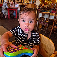 Myles is registered to the contest to win money with this photo: toddler, child, toy, remote, striped_shirt, high_chair, indoor, cafe, wooden_chair, curious, wide_eyes, person, table, people, background, lighting, decor, furniture, casual, portrait