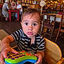 toddler, child, toy, remote, striped_shirt, high_chair, indoor, cafe, wooden_chair, curious, wide_eyes, person, table, people, background, lighting, decor, furniture, casual, portrait