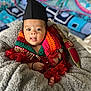 baby, infant, child, knitted_scarf, colorful_clothing, black_hat, blanket, cozy, face, expression, tongue_out, hand, foot, decorative_dagger, indoor, portrait, cute, soft_texture, patterned_background, wrapped