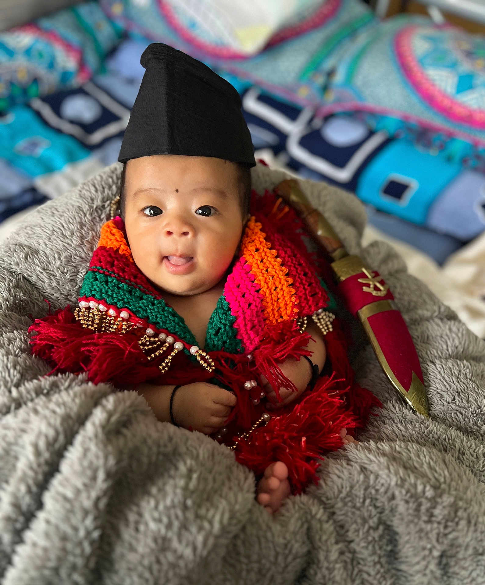 Shriyan joined the competition — help win amazing prizes! baby, infant, child, knitted_scarf, colorful_clothing, black_hat, blanket, cozy, face, expression, tongue_out, hand, foot, decorative_dagger, indoor, portrait, cute, soft_texture, patterned_background, wrapped