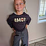 amor, braids, casual_clothing, child, confident, floor, glasses, happy, heart, indoor, natural_light, pants, person, pose, shirt, smile, sneakers, tile, wall, window