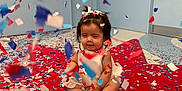 Sol is registered to the contest to win money with this photo: baby, blue, celebration, child, confetti, door, face, festive, floor, hair_bows, indoor, padded_surface, party, person, playful, red, room, sitting, wall, white