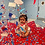 baby, blue, celebration, child, confetti, door, face, festive, floor, hair_bows, indoor, padded_surface, party, person, playful, red, room, sitting, wall, white