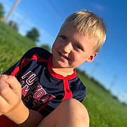 Denver is registered to the contest to win money with this photo: child, boy, smiling, outdoor, grass, blue_sky, sunlight, portrait, happy, casual_clothing, blonde_hair, nature, daylight, person, face, sitting, field, sporty_shirt, young, leisure
