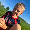 child, boy, smiling, outdoor, grass, blue_sky, sunlight, portrait, happy, casual_clothing, blonde_hair, nature, daylight, person, face, sitting, field, sporty_shirt, young, leisure