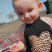 Bellamy is registered to the contest to win money with this photo: baby, child, smiling, plaid_blanket, outdoor, sunny, leopard_print, black_top, cute, portrait, face, hand, grass, playground, trash_bin, daylight, infant, person, sitting, happy