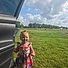 toddler, child, girl, floral_dress, car_door, grass, outdoor, sunny, sky, clouds, greenery, smiling, happy, standing, nature, daylight, young_child, park, casual, portrait