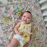 Amina joined the competition — help win amazing prizes! baby, infant, smiling, crib, floral_pattern, toy, elephant_toy, yellow, white_clothing, lying_down, happy, cute, child, playful, indoors, colorful, flower_print, skin, cheerful, one_leg_up