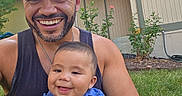 Elijah is registered to the contest to win money with this photo: man, baby, smiling, outdoor, grass, greenery, house, tank_top, cap, beard, happy, child, portrait, daylight, plants, lawn, casual_clothing, family, parent, baby_shirt